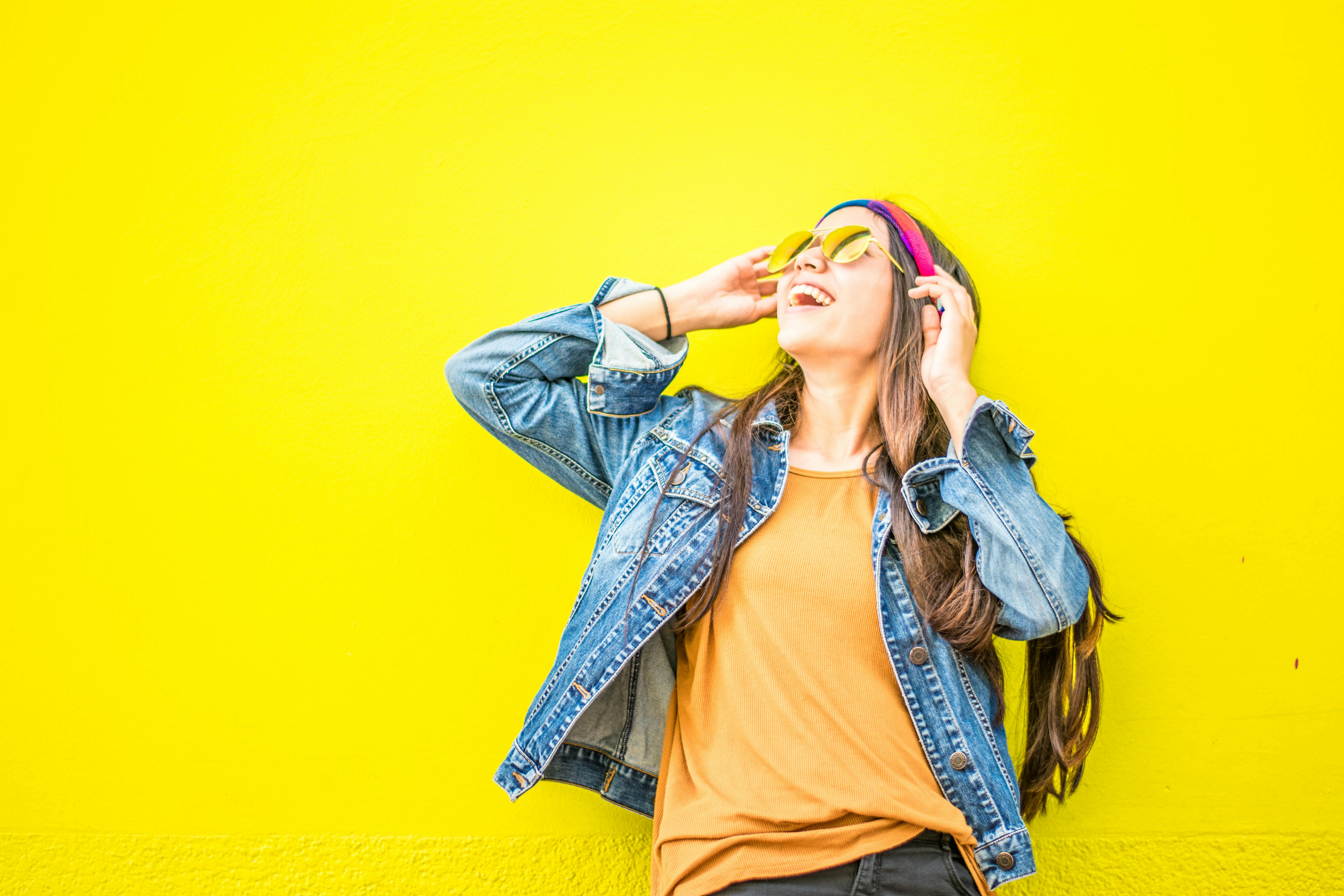 Smiling woman in sunglasses stands against vibrant yellow wall, radiating happiness.