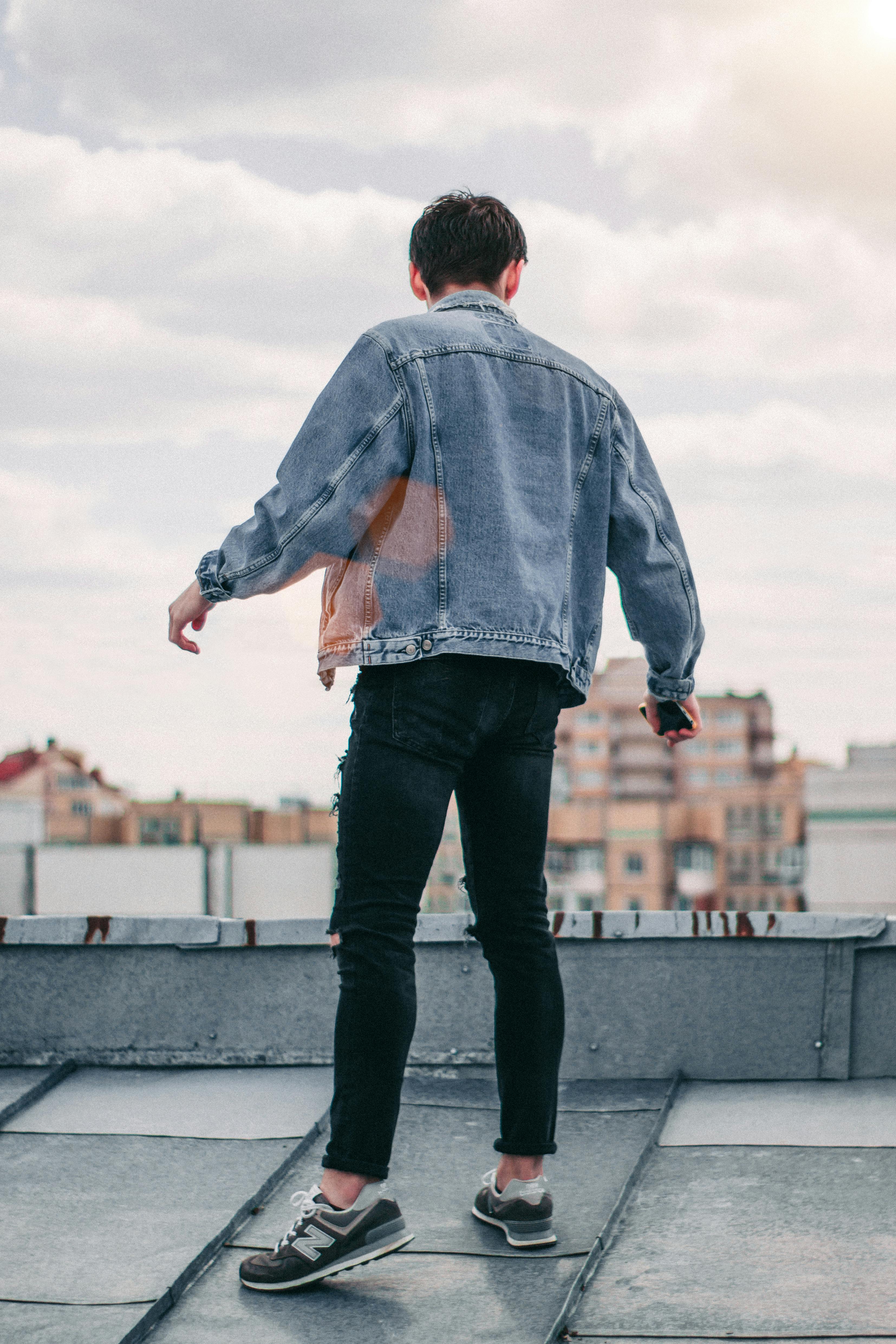 Fashionable young man wearing a denim jacket standing on a rooftop with cloudy skies.