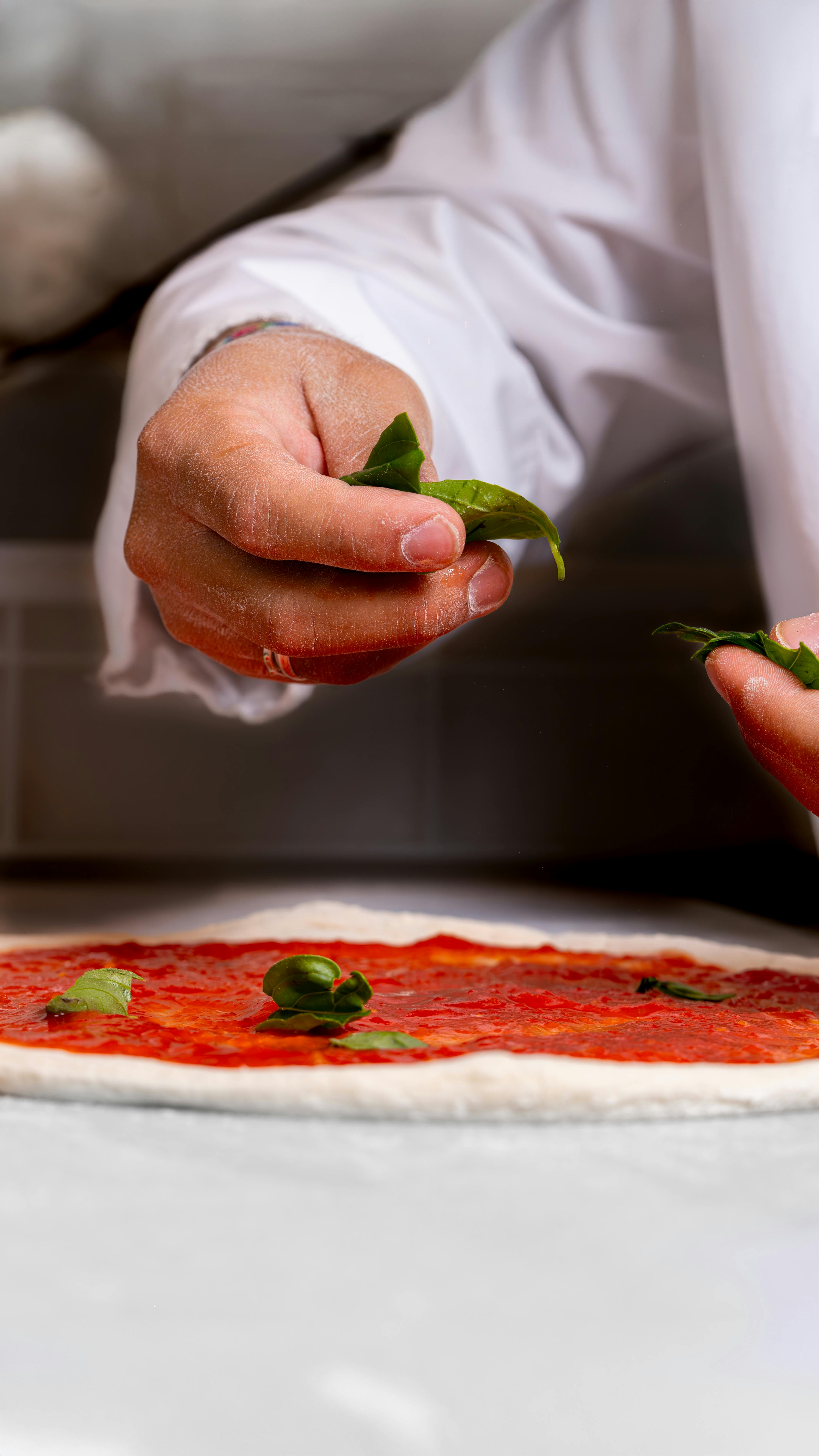 Close-up of hands adding basil on pizza dough with tomato sauce.