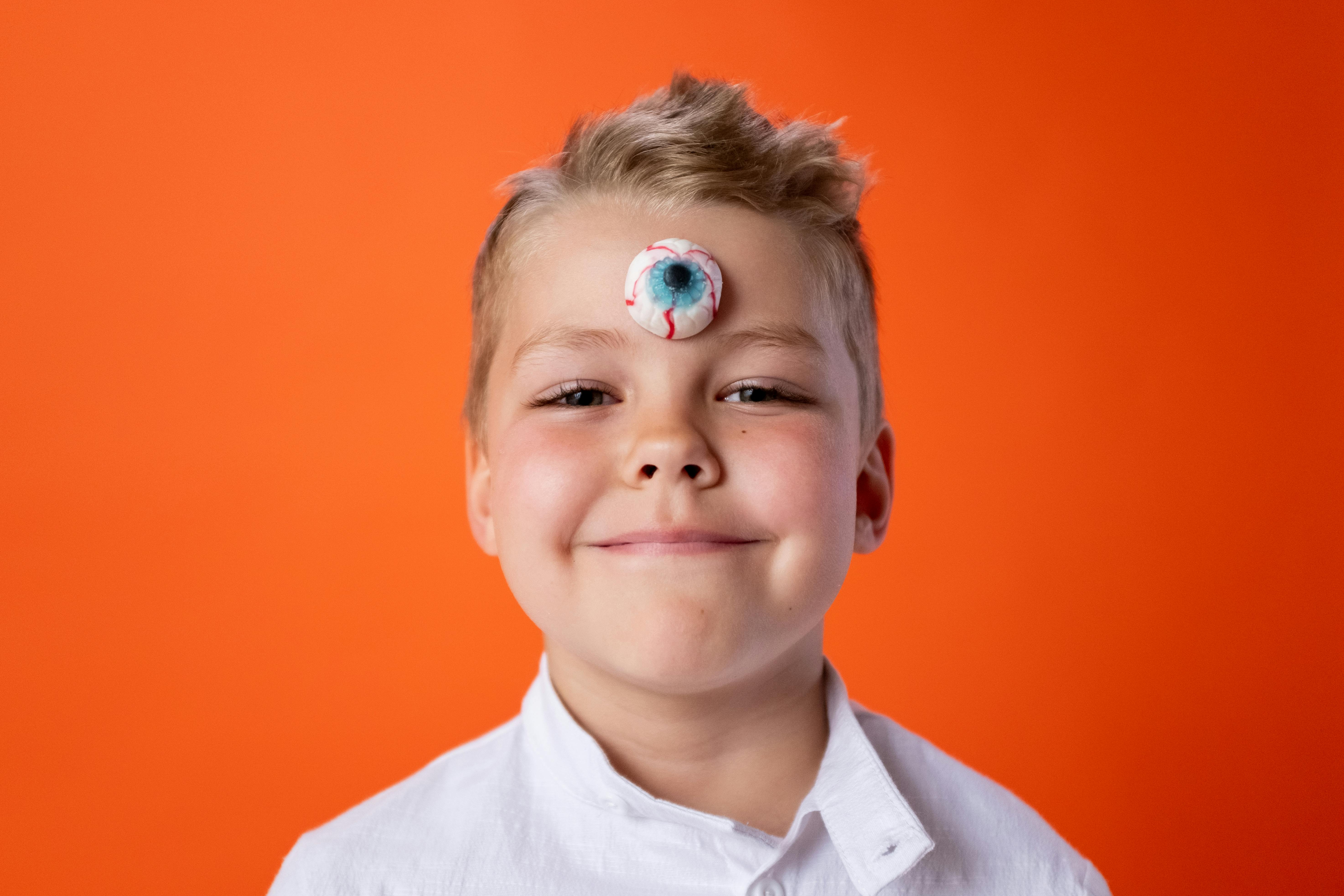 Smiling child with candy eyeball on forehead against orange background, showcasing creativity and fun.