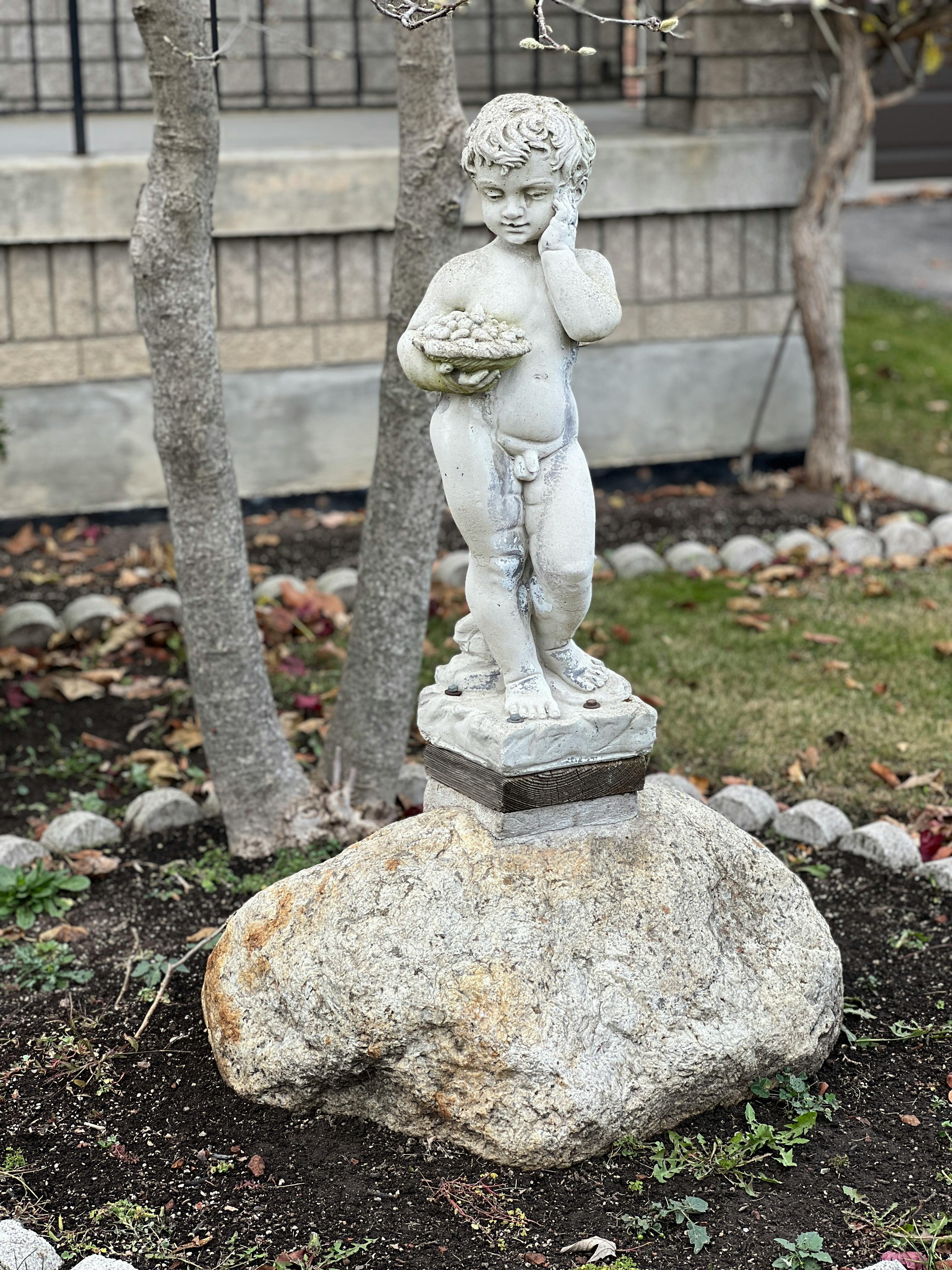 Serene garden scene with a child statue perched on a stone, surrounded by greenery.