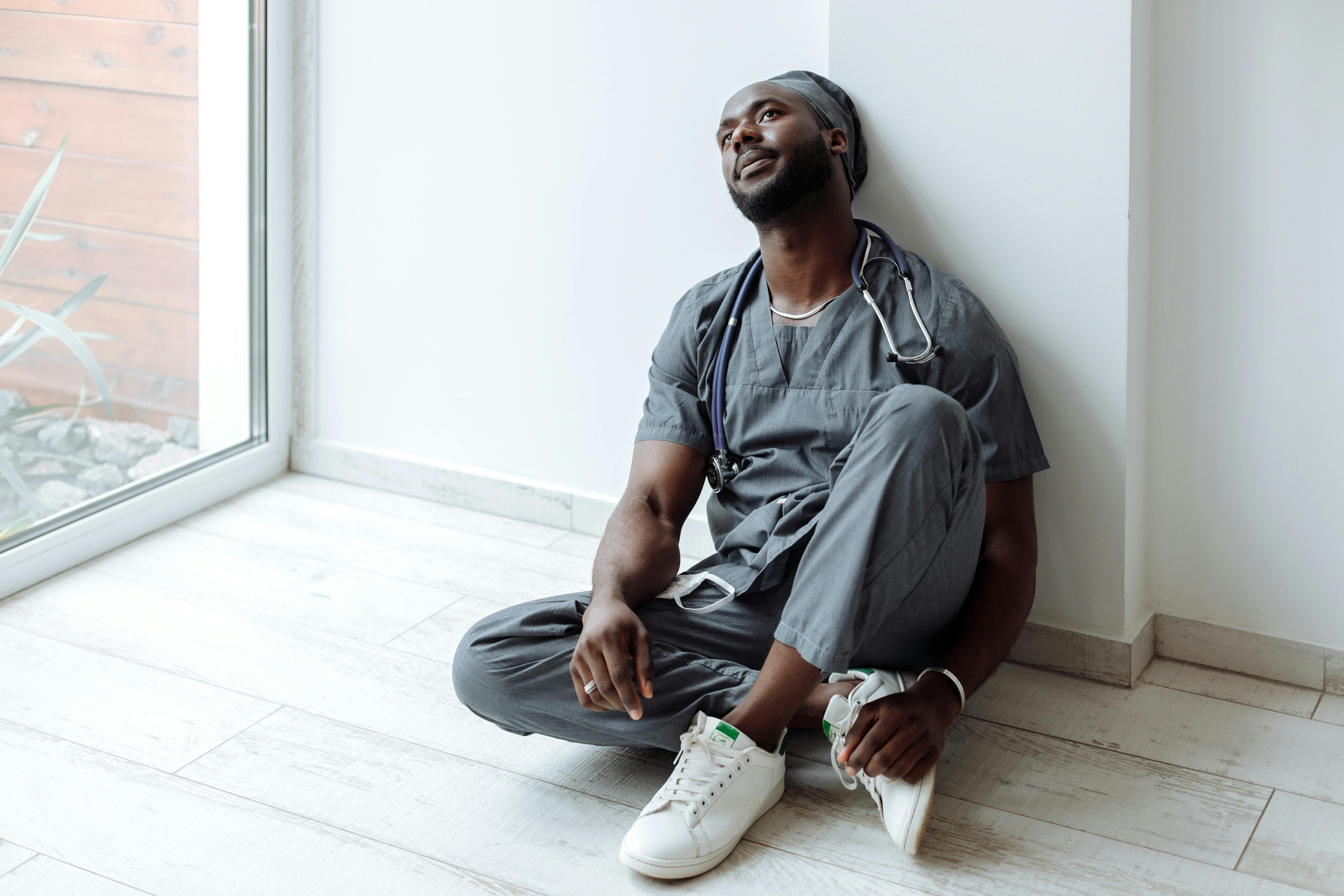 A tired healthcare professional in gray scrubs resting against a wall, reflecting exhaustion.