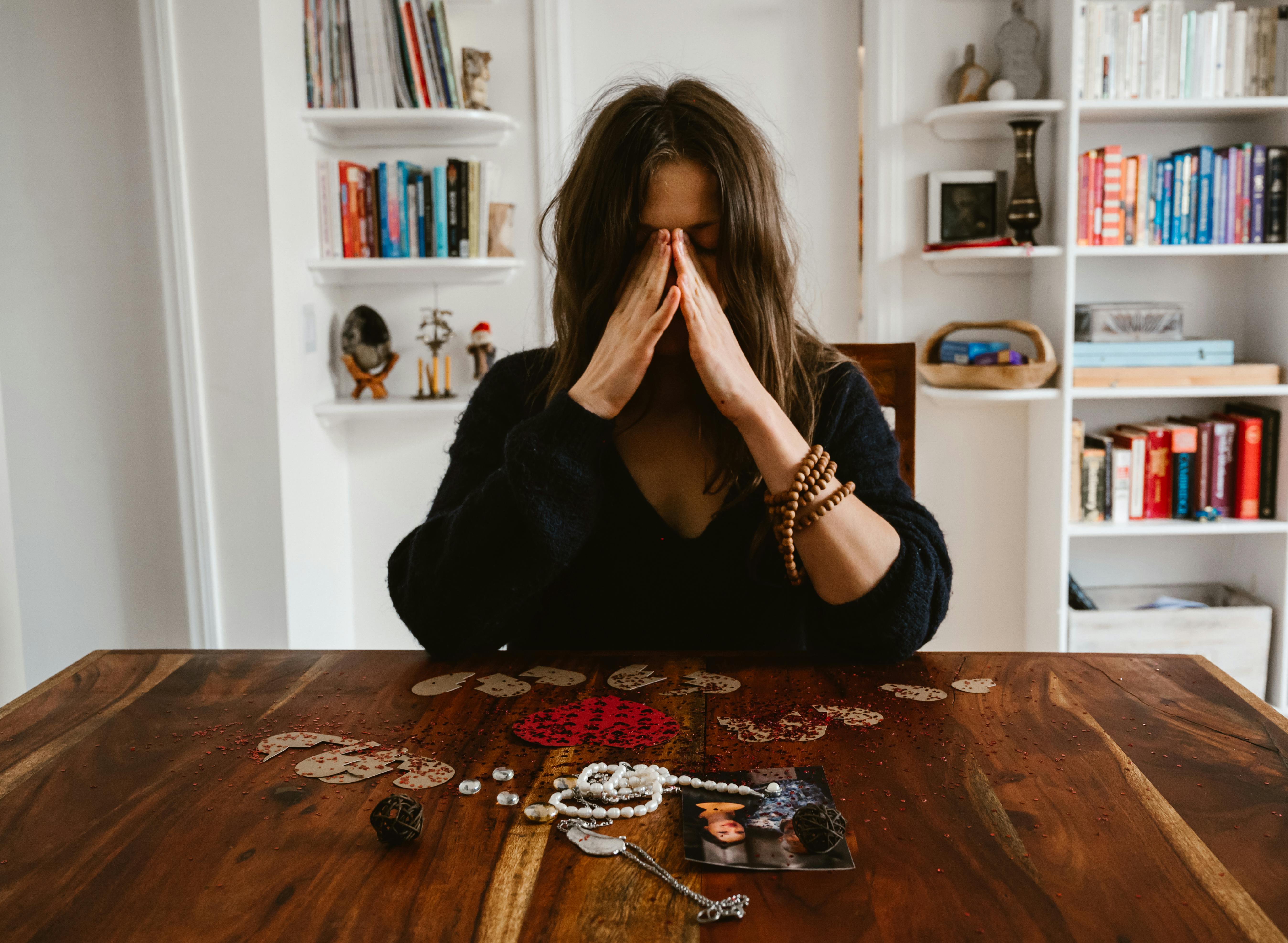 A woman in deep thought sits at a table with beads and a photo, conveying introspection.