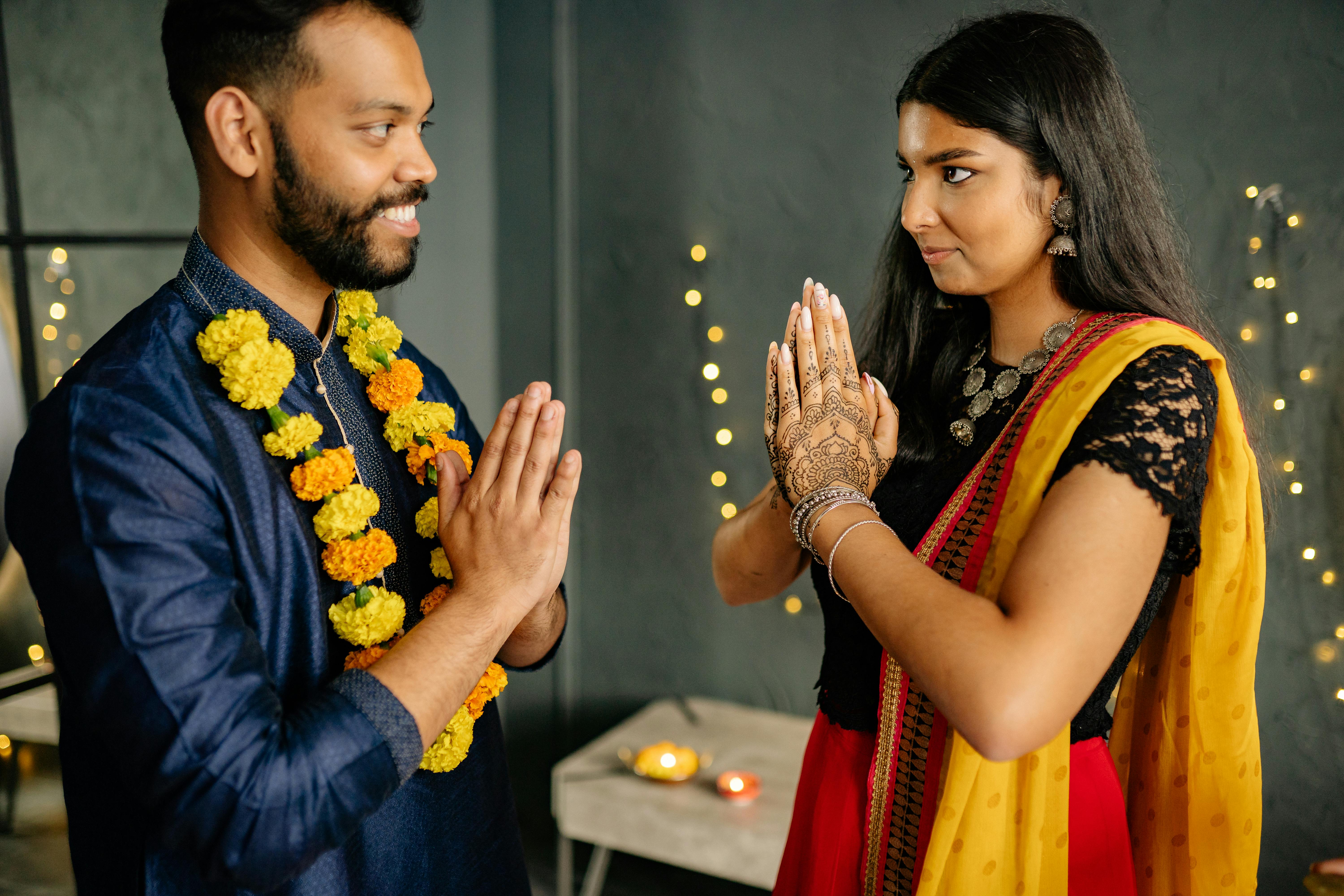 An Indian couple in traditional attire exchanging a warm namaste greeting indoors.