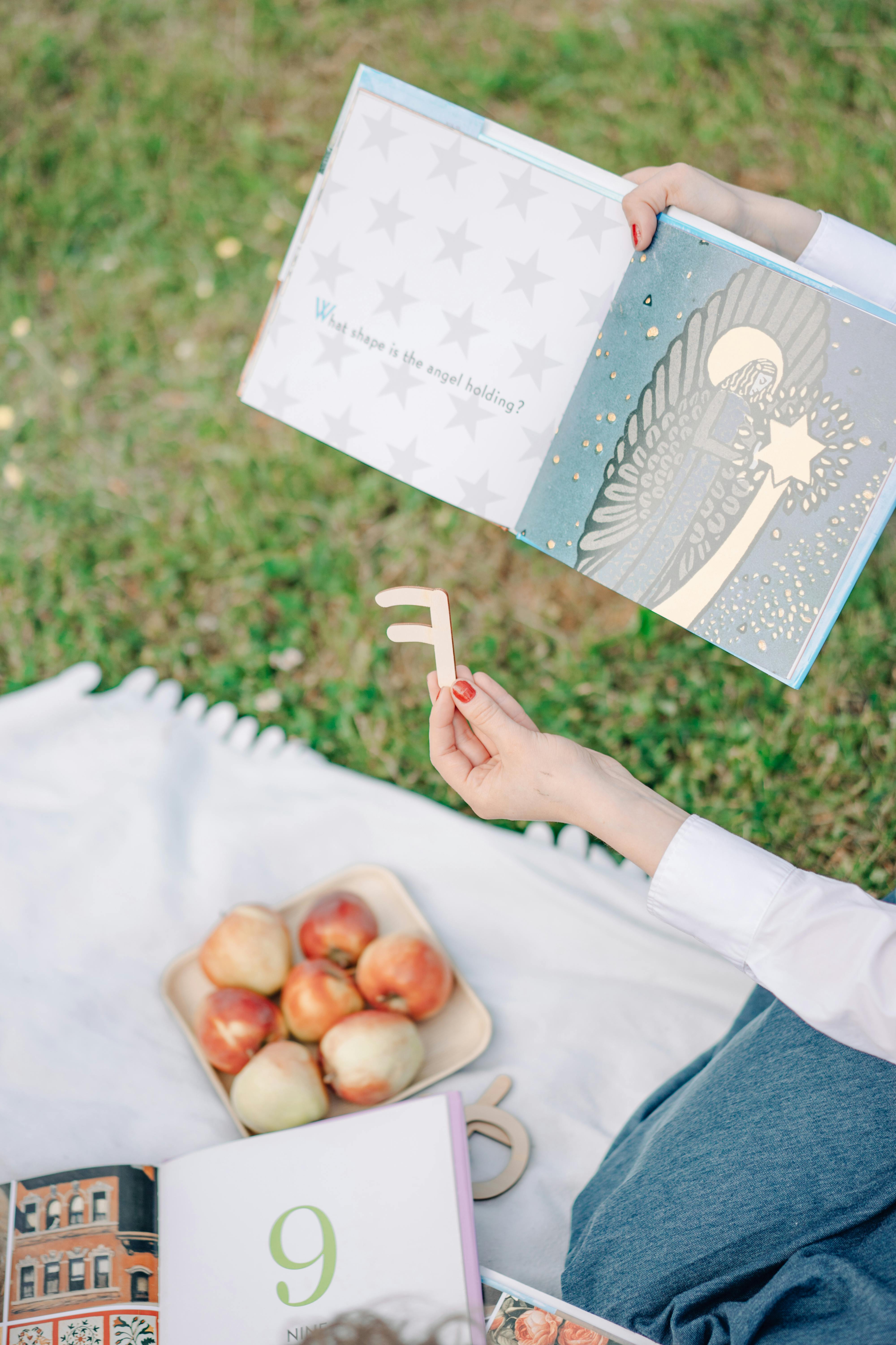 A child reads a book outdoors with apples on a picnic blanket, enjoying a sunny day.